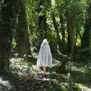 May include: A white hooded cloak made of fabric that drapes down to the mid-thigh, featuring a pointed hood and a loose, flowing silhouette. The garment is worn over bare legs and is captured from behind as it moves through a wooded area.
