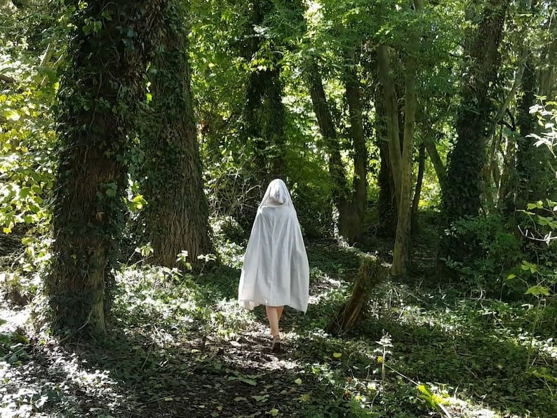 May include: A white hooded cloak made of fabric that drapes down to the mid-thigh, featuring a pointed hood and a loose, flowing silhouette. The garment is worn over bare legs and is captured from behind as it moves through a wooded area.