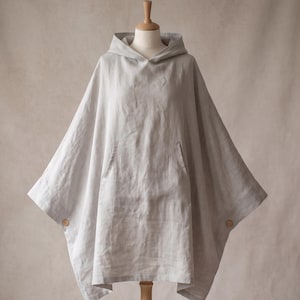 May include: A light gray hooded poncho featuring a large front kangaroo pocket and two small wooden buttons on the side seams. The garment has a loose, draped silhouette and is displayed on a mannequin, showcasing its textured fabric construction.