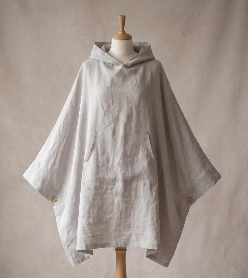 May include: A light gray hooded poncho featuring a large front kangaroo pocket and two small wooden buttons on the side seams. The garment has a loose, draped silhouette and is displayed on a mannequin, showcasing its textured fabric construction.