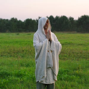 May include: A beige hooded cloak with long, wide sleeves and two visible wooden buttons at the chest. The garment is worn over a matching beige tunic and grey trousers, featuring a loose, draped fit that extends to the mid-thigh area.