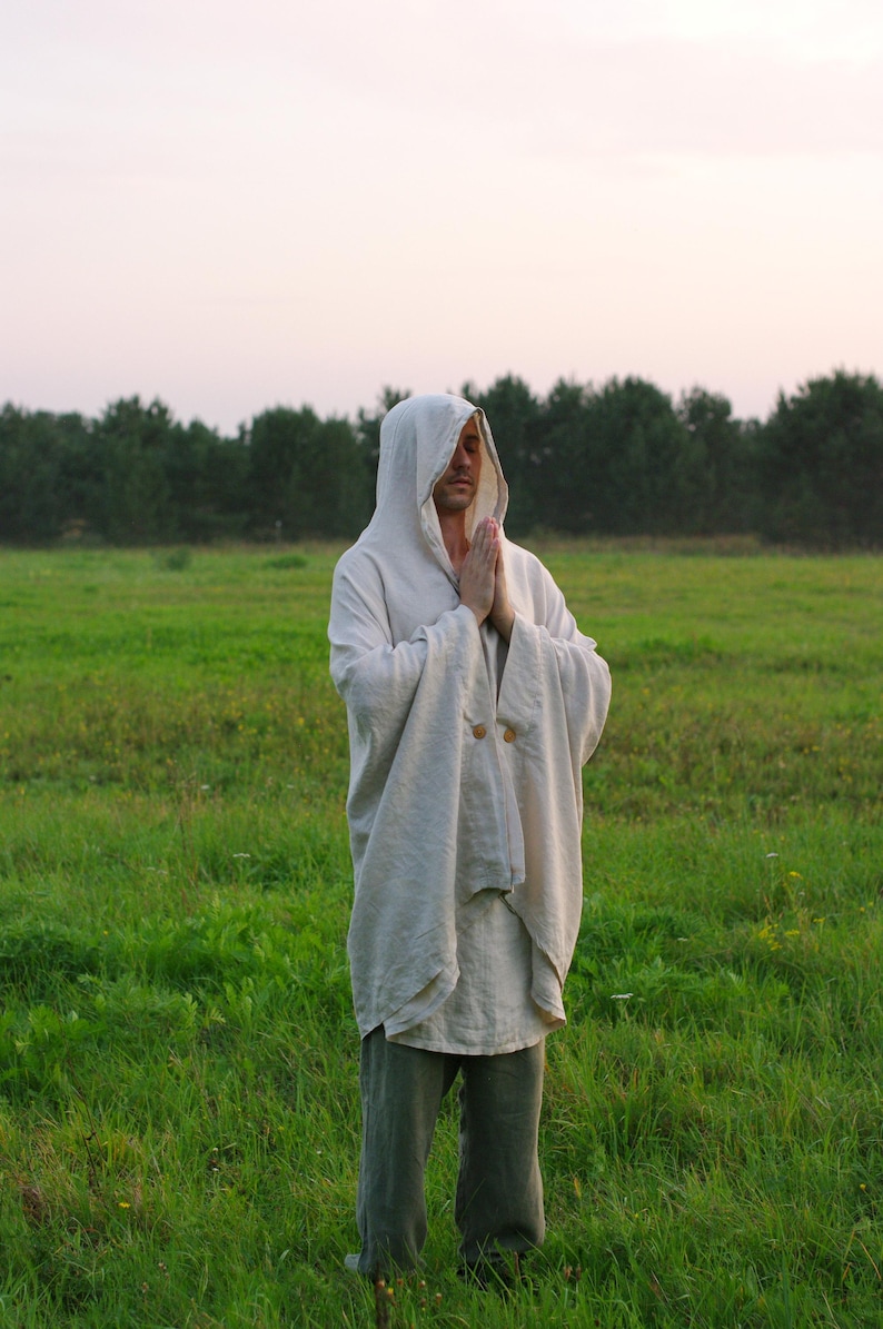 May include: A beige hooded cloak with long, wide sleeves and two visible wooden buttons at the chest. The garment is worn over a matching beige tunic and grey trousers, featuring a loose, draped fit that extends to the mid-thigh area.