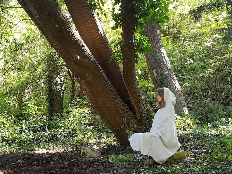 May include: A light beige hooded poncho featuring a loose, draped silhouette with long sleeves and a soft, textured fabric finish. The garment covers the upper body and extends down to the knees, providing a relaxed fit with a simple hood.