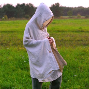 May include: An off-white hooded poncho made of a textured fabric, featuring a single visible wooden button closure on the side. The garment has a loose, draped fit with wide sleeves and a deep hood, worn over dark gray trousers and dark shoes.