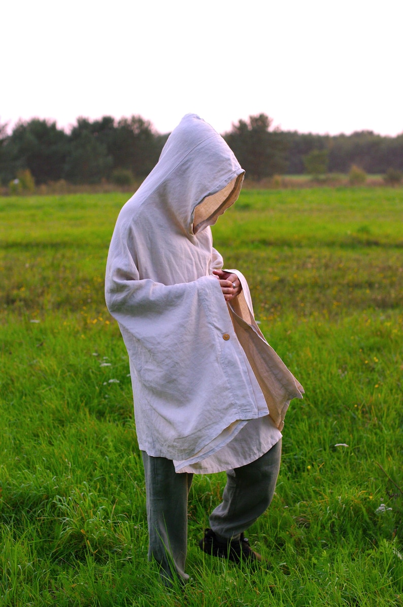 May include: An off-white hooded poncho made of a textured fabric, featuring a single visible wooden button closure on the side. The garment has a loose, draped fit with wide sleeves and a deep hood, worn over dark gray trousers and dark shoes.