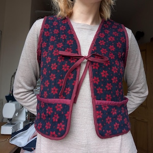 May include: A sleeveless waistcoat featuring a dark navy blue base with a repeating red floral pattern. The garment has a maroon fabric trim along the edges, two front pockets, and a tie closure at the centre of the chest made from matching fabric.