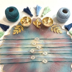 May include: Seven thin string bracelets featuring small circular silver-toned charms with an Om symbol. The bracelets are arranged on a round, scalloped-edge resin tray with a marbled blue, pink, and gold finish, accompanied by yarn and tassels.