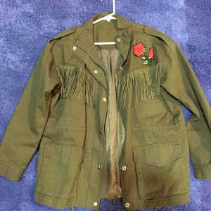 May include: A green utility jacket with a rose embroidered on the right side. The jacket has a fringe trim around the bottom and a drawstring at the waist.