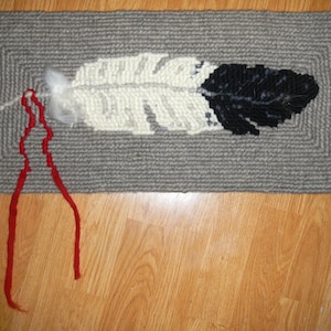 Könnte beinhalten: Ein grauer Teppich mit einem weißen und schwarzen Federdesign. Der Teppich hat einen roten Fransen an den Rändern.