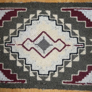 May include: A hand-woven rug with a geometric pattern in shades of gray, white, and burgundy. The rug has a central diamond shape with a smaller diamond inside. The rug is made of wool and has a fringed edge.