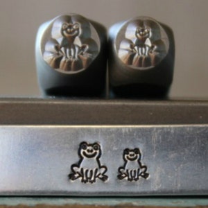 May include: Two silver metal stamps with a frog design. The stamps are sitting on a silver metal surface with two smaller frog designs stamped into the surface.