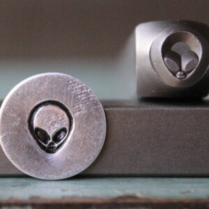May include: A close-up of a metal stamp with an alien head design. The stamp is silver and has a circular shape. The alien head is black and has large eyes and a small mouth.