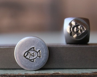 May include: Two metal stamps with a fish design. One is a small, round stamp with a fish outline. The other is a cube-shaped stamp with a fish carved into the side. The stamps are silver and dark gray.
