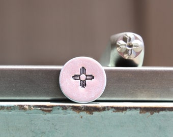 May include: Close-up of a silver metal stamp with a design of the Zia symbol, a traditional symbol of the Pueblo people of New Mexico. The stamp is on a gray surface.
