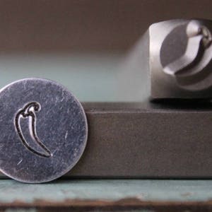 Puede incluir: Un sello de metal con un diseño de chile. El sello se utiliza para crear impresiones en metal.