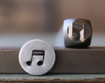 May include: A metal stamp and an impression of a musical note. The stamp is a dark gray cube with the musical note symbol on one face. The impression is a small, round, silver-colored disc with the musical note symbol in black.