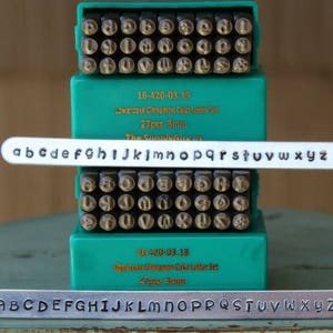 May include: Two sets of metal letter stamps in a green plastic case. The top set is lowercase letters and the bottom set is uppercase letters. Each set has 27 letters and is 3mm in size. The text on the case reads "Lowercase Cinnamon Cake Letter Set 27pcs 3mm The SunnyGum us" and "Uppercase Cinnamon Cake Letter Set 27pcs 3mm".