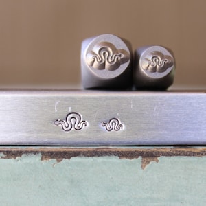 May include: Two metal stamps with a snake design. The stamps are on a silver metal surface. The snake design is also stamped on the surface.