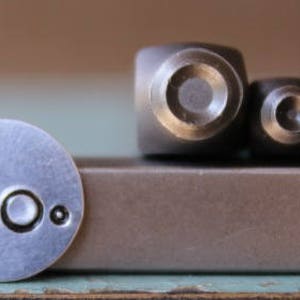 May include: Three metal stamp tools with different sized circular designs. The stamps are used to create impressions in metal. The stamps are on a brown surface.