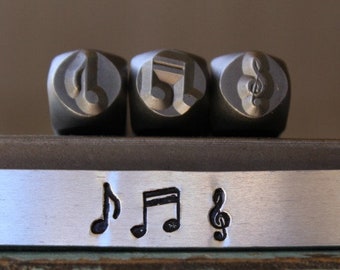 May include: Three metal stamp blocks with musical note designs. The blocks are silver and have a slightly textured surface. The notes are a treble clef, a quarter note, and a half note.
