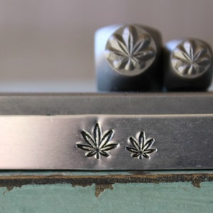 May include: A silver metal bar with two small leaf designs stamped into the surface. The bar is sitting on top of a green surface.