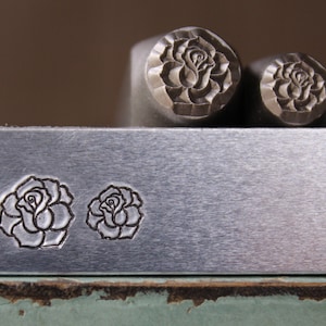 Könnte beinhalten: Zwei Metallstempel mit Rosenmotiven. Die Stempel sind auf einer Metalloberfläche mit zwei Abdrücken des Rosenmotivs abgebildet.