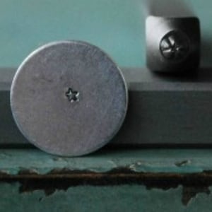 May include: A close-up of a silver metal stamp with a five-pointed star design. The stamp is used to create impressions in metal.
