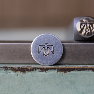 May include: Two metal stamps with a stylized bird design. The stamp on the left is a round metal stamp with a silver finish. The stamp on the right is a rectangular metal stamp with a silver finish.