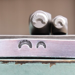 Op de afbeelding: Twee metalen stempels met een waaierontwerp, naast een afdruk van hetzelfde ontwerp op een metalen oppervlak. De stempels zijn cilindrisch met het waaierontwerp aan het platte uiteinde. De afdruk is een reeks gebogen lijnen.