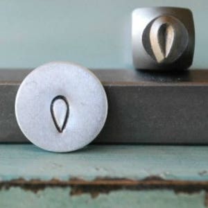 Könnte beinhalten: Ein Stempel aus silbernem Metall mit einem tropfenförmigen Design. Der Stempel wird verwendet, um Abdrücke in Metall zu erstellen.