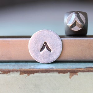 May include: A close-up of a metal stamp with a pointed arrow design. The stamp is used to create impressions on metal.