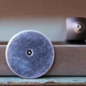 May include: Close-up of a silver-colored metal stylus tip with a small, round indentation in the center. The tip is resting on a brown surface.