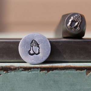 May include: A close-up of two metal stamps. The stamp on the left is a round stamp with a design of two hands clasped in prayer. The stamp on the right is a rectangular stamp with a design of two hands clasped in prayer.