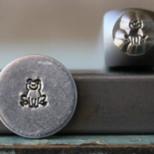 May include: A close-up of a metal stamp with a frog design. The stamp is used to create impressions in metal.