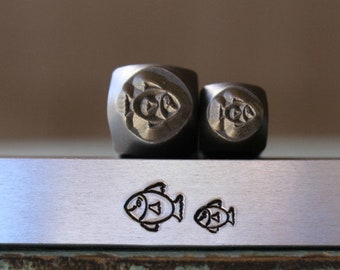 May include: Two metal stamps with fish designs. The larger stamp has a design of three fish, while the smaller stamp has a design of two fish. The stamps are sitting on a metal bar with two fish designs stamped on it.