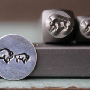 May include: Two metal stamp heads with a design of a bison, one larger and one smaller. The stamp heads are on a metal bar. A silver metal disc with the same bison design is also shown.