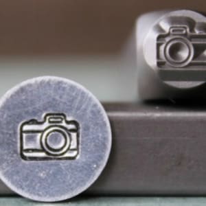 May include: A silver metal stamp with a camera design. The stamp is round and has a raised design of a camera with a lens and a shutter.