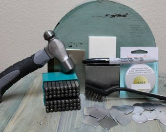 Könnte beinhalten: Ein Satz Metallstempel in einem türkisfarbenen Kunststoffkoffer, ein Hammer, eine Metallbürste, ein silberner Stift, eine Rolle Klebeband und verschiedene Metallscheiben. Das Klebeband trägt die Aufschrift "Peace" und "Steel Straight Tape".