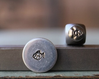May include: Two metal stamps with a fish design. One stamp is a small, round disc with the fish etched into the surface. The other stamp is a small, square block with the fish design on one side. The stamps are silver in color.