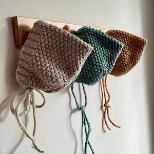 Puede incluir: Tres gorros de bebé de punto en beige, verde azulado y amarillo mostaza cuelgan de un perchero de madera. Cada gorro tiene un patrón de punto texturizado y lazos. Los gorros se exhiben contra una pared blanca.