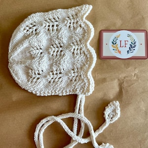 Può includere: Un berretto per neonato in crochet bianco con un bordo a festoni e un lungo laccio. Il berretto ha un delicato motivo a pizzo ed è legato con due piccole foglie all'uncinetto.