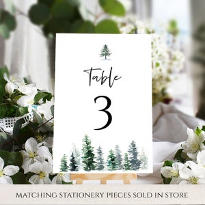 May include: White table number card with the text "Table 3" in black script font. A small watercolor illustration of a pine tree is at the top of the card. A row of watercolor pine trees are at the bottom of the card.