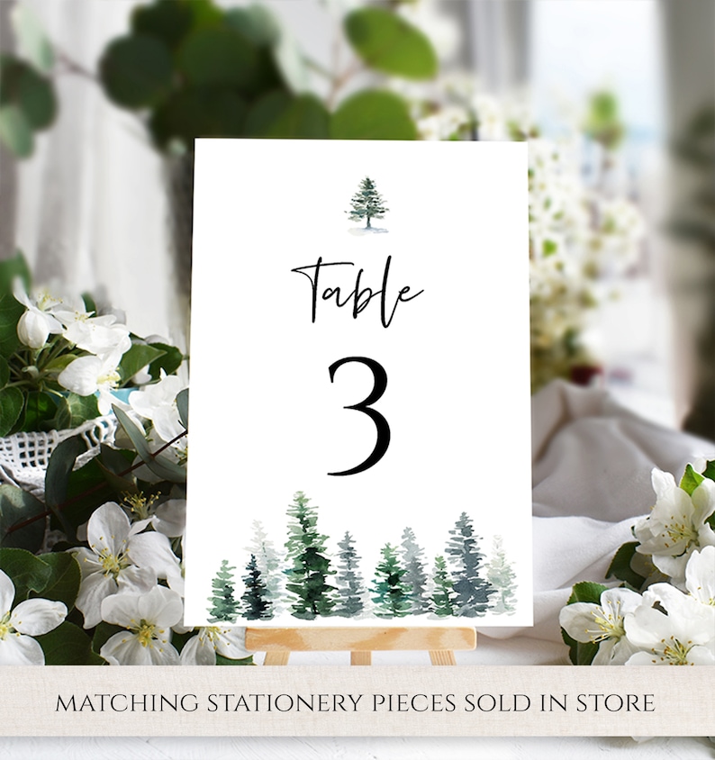 May include: White table number card with the text "Table 3" in black script font. A small watercolor illustration of a pine tree is at the top of the card. A row of watercolor pine trees are at the bottom of the card.