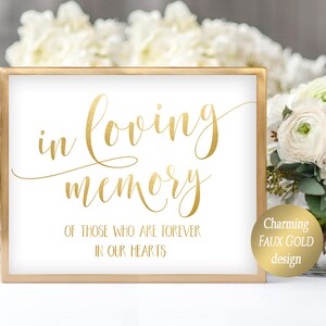 May include: A white sign with a gold frame and gold lettering that reads "in loving memory of those who are forever in our hearts". The sign is sitting on a white table with a bouquet of white and pink flowers in the background.