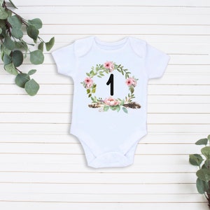 May include: White baby onesie with a floral wreath design and the number "1" in the center. The wreath is made of pink flowers, green leaves, and brown feathers.