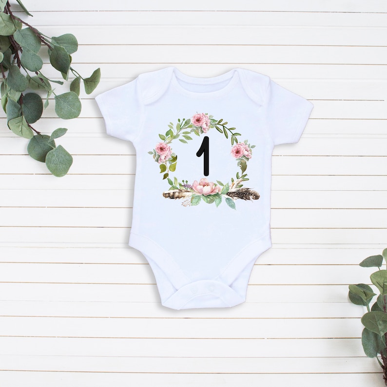 May include: White baby onesie with a floral wreath design and the number "1" in the center. The wreath is made of pink flowers, green leaves, and brown feathers.