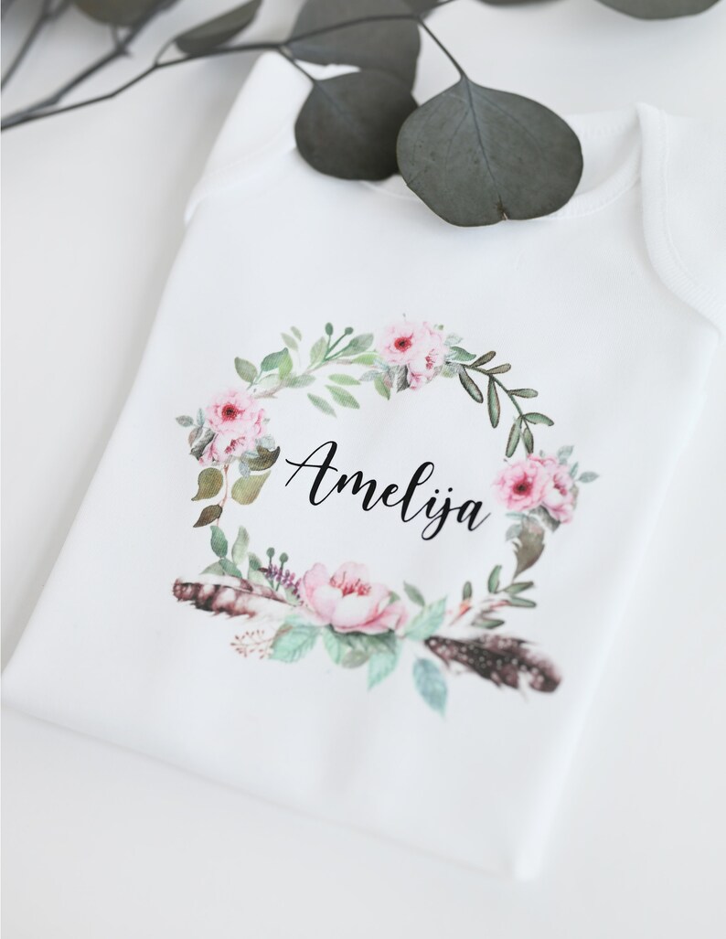 May include: White baby onesie with a floral wreath design and the name Amelia printed in black.