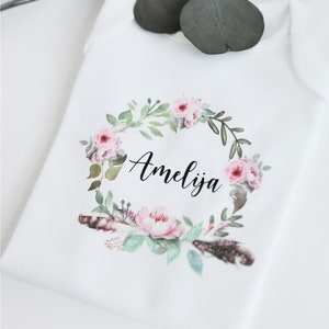 K&ouml;nnte beinhalten: Wei&szlig;er Baby-Strampler mit einem floralen Kranz-Design und dem Namen Amelia in Schwarz gedruckt.