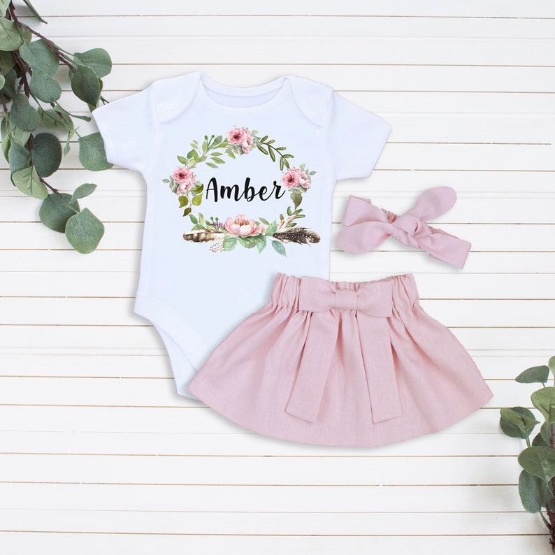 May include: A baby outfit set with a white bodysuit featuring a floral wreath with the name "Amber" in the center, a pink skirt with a bow, and a pink headband with a bow.