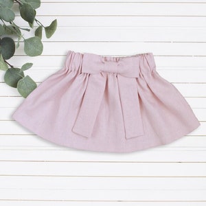 May include: A pale pink skirt with a large bow at the waist. The skirt is made of a soft, lightweight fabric.